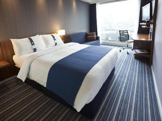 Cozy hotel room with a plush king-sized bed, navy blue accents, and a functional workspace, featuring a panoramic cityscape visible through the window